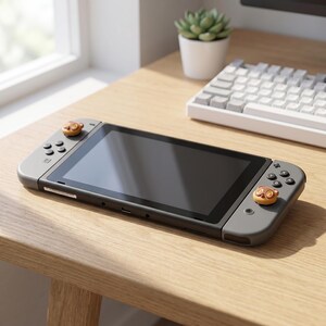 Op de afbeelding: Een grijze Nintendo Switch-console met aangepaste knoppen met een cartoon-dierontwerp. De console staat op een houten oppervlak, in de buurt van een raam en een wit toetsenbord.