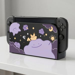 Op de afbeelding: Zwart Nintendo Switch-dock met een paars en zwart ontwerp met stripfiguren, sterren en wolken. Het dock heeft een paars personage aan de onderkant en een geel personage aan de bovenkant.