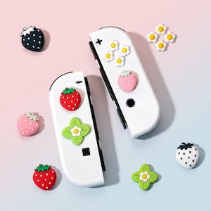 Puede incluir: Dos mandos de juego blancos decorados con pequeños dijes de silicona con forma de fresas rojas, fresas rosas, margaritas blancas y tréboles de cuatro hojas verdes. Los dijes están dispuestos sobre la superficie de los mandos.