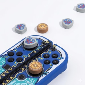 Könnte beinhalten: Blau-weißer Nintendo Switch Joy-Con-Controller mit passenden Daumengriffen. Die Daumengriffe weisen ein Schilddesign mit roten und blauen Akzenten auf. Zusätzliche braune Daumengriffe mit goldenem Design sind ebenfalls vorhanden.