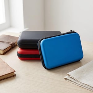 May include: Three small, rectangular zippered cases in black, red, and blue are stacked on a light wood surface. A blue case is in the foreground, with a black zipper and a smooth, glossy finish. The cases appear to be for storing small electronics or accessories.