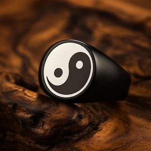May include: A black ring with a polished finish, featuring a Yin and Yang symbol in silver and black. The ring is set against a wooden background, highlighting the contrast of the colors and textures. The ring is a signet style.