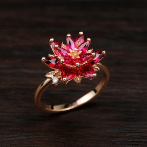 May include: A gold ring with a flower design, featuring red gemstone petals and a small diamond. The ring's band is delicate, and the flower is the central element, highlighting the intricate details of the petals.