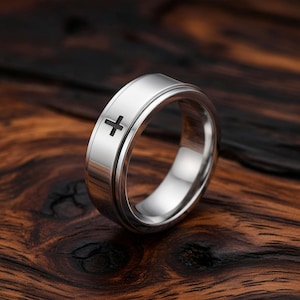 Könnte beinhalten: Ein silberner Ring mit einem schwarzen Kreuzsymbol. Der Ring hat eine glatte, polierte Oberfläche und ein drehbares äußeres Band. Das Kreuz ist in das Band geätzt und fügt dem Design ein subtiles Detail hinzu.
