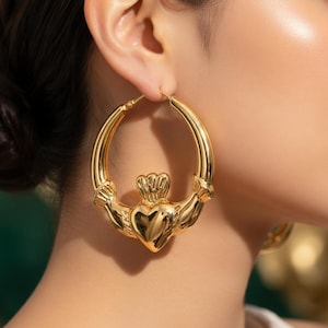 Puede incluir: Pendientes de aro dorados con diseño Claddagh. Los pendientes tienen un corazón en el centro, coronado y abrazado por dos manos. Los aros son lisos y pulidos, reflejando la luz.