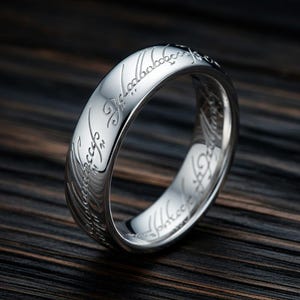 May include: A polished silver ring with engraved lettering. The ring's surface reflects light, and the engraved text is visible around the band. The ring is set against a dark wood grain background.