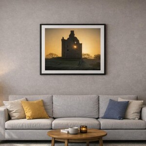 May include: A framed print of a castle silhouette against a sunset. The artwork is displayed above a gray sofa with colorful throw pillows. A side table with a lamp and a coffee table with decor complete the living room setting.