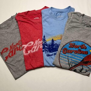 K&ouml;nnte beinhalten: Vier gefaltete T-Shirts in Grau, Rot und Blau. Die Shirts zeigen North Carolina-Grafiken, darunter einen Kardinal, eine Berglandschaft und Schrifttext. Die Shirts sind aus einem weichen Material.