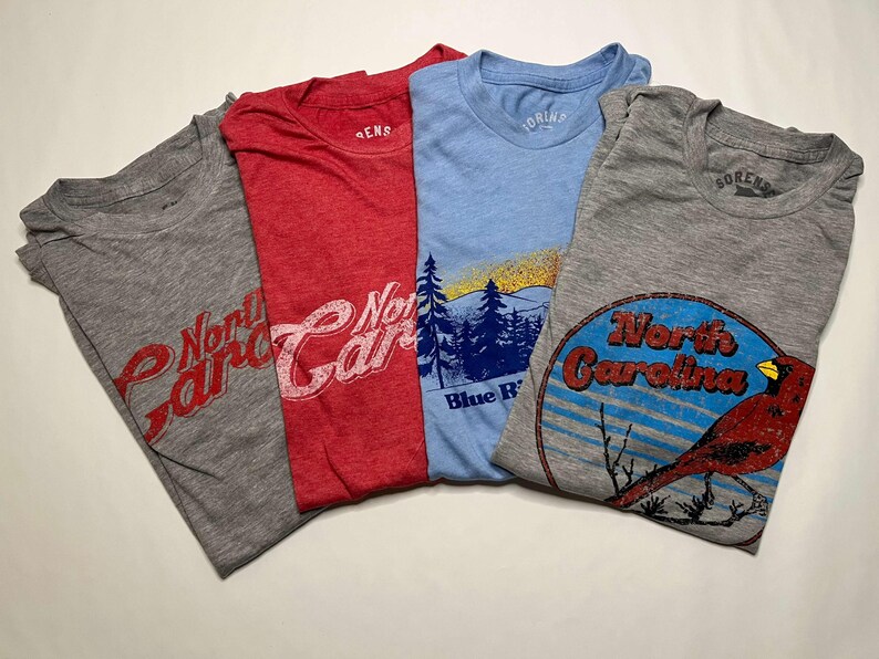 K&ouml;nnte beinhalten: Vier gefaltete T-Shirts in Grau, Rot und Blau. Die Shirts zeigen North Carolina-Grafiken, darunter einen Kardinal, eine Berglandschaft und Schrifttext. Die Shirts sind aus einem weichen Material.