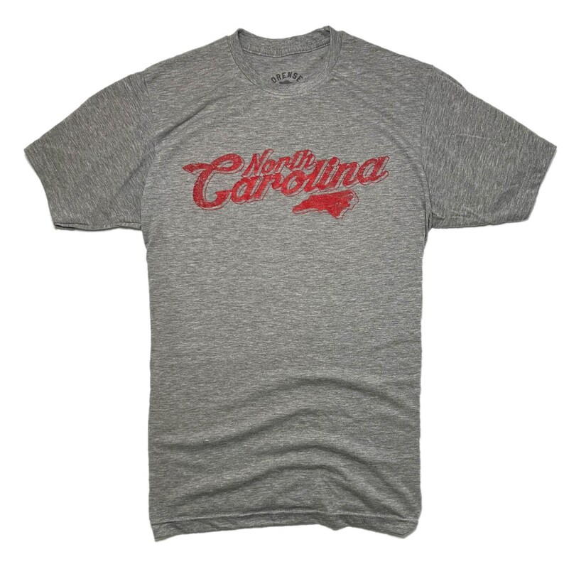 K&ouml;nnte beinhalten: Ein meliertes graues T-Shirt mit dem Text "North Carolina" in verblichener roter Schrift und einer roten Umrisszeichnung des Bundesstaates. Das Shirt besteht aus einem weichen, bequemen Material.