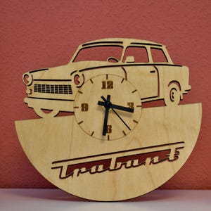 Könnte beinhalten: Wanduhr aus Holz mit einer Autosilhouette und einem Zifferblatt. Die Uhr hat eine natürliche Holzoberfläche mit schwarzen Zeigern und Ziffern. Das Wort "Trabant" ist unten ausgeschnitten. Die Uhr ist ein dekorativer Gegenstand.