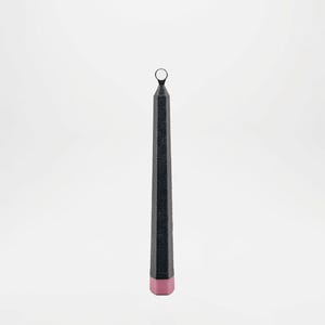 Puede incluir: Una vela alta y negra con una superficie texturizada y un pequeño anillo en la parte superior. La base de la vela es de color rosa suave. La vela está sobre un fondo blanco liso.