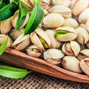 May include: Close-up of a wooden bowl filled with pistachios. The nuts are in their shells, some cracked open to reveal the green nut inside. Green leaves are in the foreground, adding a natural element to the image.