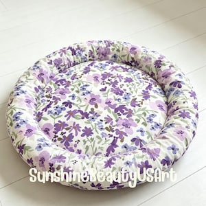 May include: A round, cushioned pet bed featuring a white fabric base with a repeating floral pattern in shades of purple, lavender, and light green. The text SunshineBeautyUSArt is printed in white across the bottom of the circular bed.