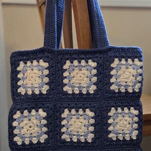 May include: A square crochet tote bag constructed from nine individual granny squares. The squares feature a central pattern of white and light blue yarn, bordered by a solid dark blue yarn. The bag includes two matching dark blue crochet handles.
