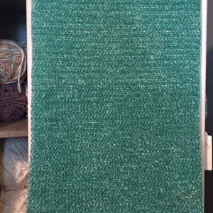 May include: A rectangular, teal-colored, plush blanket. The blanket is made of a soft, textured material and is displayed on a white rack. Various balls of yarn in shades of pink, cream, and purple are visible on a shelf in the background.
