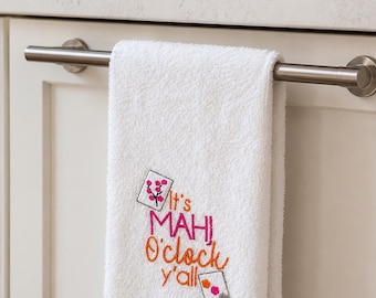 May include: White hand towel with embroidered text "It's Mahi O'clock y'all" in pink and orange. Two small embroidered card-like designs with floral and dot patterns are also present. The towel is hanging on a silver bar.