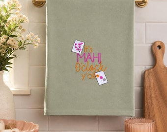 May include: A sage green hand towel featuring embroidered text that reads It's MAHI O'clock y'all in pink and orange thread. The design includes two small white rectangular cards, one with a pink floral branch and one with colourful flowers.