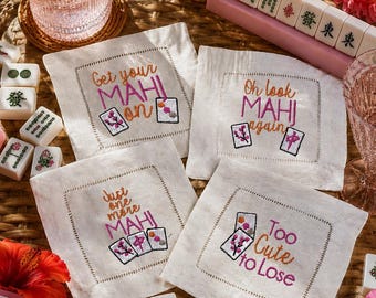 May include: Four square off-white fabric napkins featuring embroidered text and Mahjong tile illustrations. The text reads Get your MAHI on, Oh look MAHI again, Just one more MAHI, and Too cute to lose, accented with pink and orange thread.