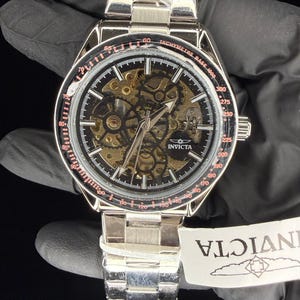 May include: Silver-toned Invicta watch with a black face and visible internal gears. The watch features a tachymeter bezel with red accents and a silver link band. The watch is new, with a tag attached.