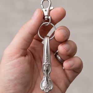 May include: A silver-coloured metal keyring featuring a decorative handle from a piece of cutlery. The handle has an embossed floral pattern and is attached to a circular split ring and a lobster claw clasp, creating a functional accessory.