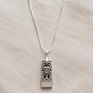 Peut inclure: Un collier pendentif rectangulaire de couleur argentée présentant un motif floral et des volutes en relief sur la partie supérieure, suspendu à une fine chaîne carrée. La partie inférieure du pendentif est en métal argenté lisse et poli.