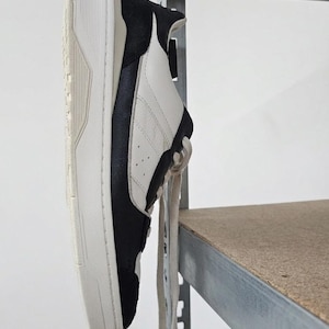 Könnte beinhalten: Ein schwarz-weißer Sneaker mit einer dicken, cremefarbenen Sohle. Der Schuh hat schwarze Wildleder-Einsätze und weiße Lederteile. Die Schnürsenkel sind hellbeige. Der Sneaker wird auf einem Metallregal präsentiert.