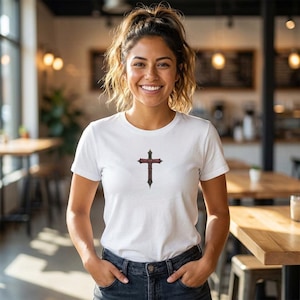 Könnte beinhalten: Ein weißes T-Shirt mit einem Kreuzdesign in Rot und Schwarz. Das Kreuz befindet sich mittig auf der Brust. Das Shirt hat kurze Ärmel und ist aus weichem Material. Die Person trägt eine dunkelblaue Jeans.