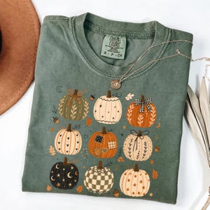 May include: A sage green short-sleeve t-shirt featuring a graphic print of nine decorative pumpkins in various patterns, including plaid, stars, and floral motifs. The shirt has a crew neckline and a Comfort Colors brand label at the inner collar.