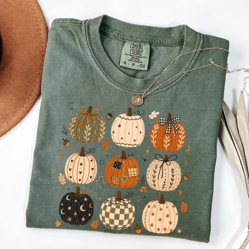 May include: A sage green short-sleeve t-shirt featuring a graphic print of nine decorative pumpkins in various patterns, including plaid, stars, and floral motifs. The shirt has a crew neckline and a Comfort Colors brand label at the inner collar.