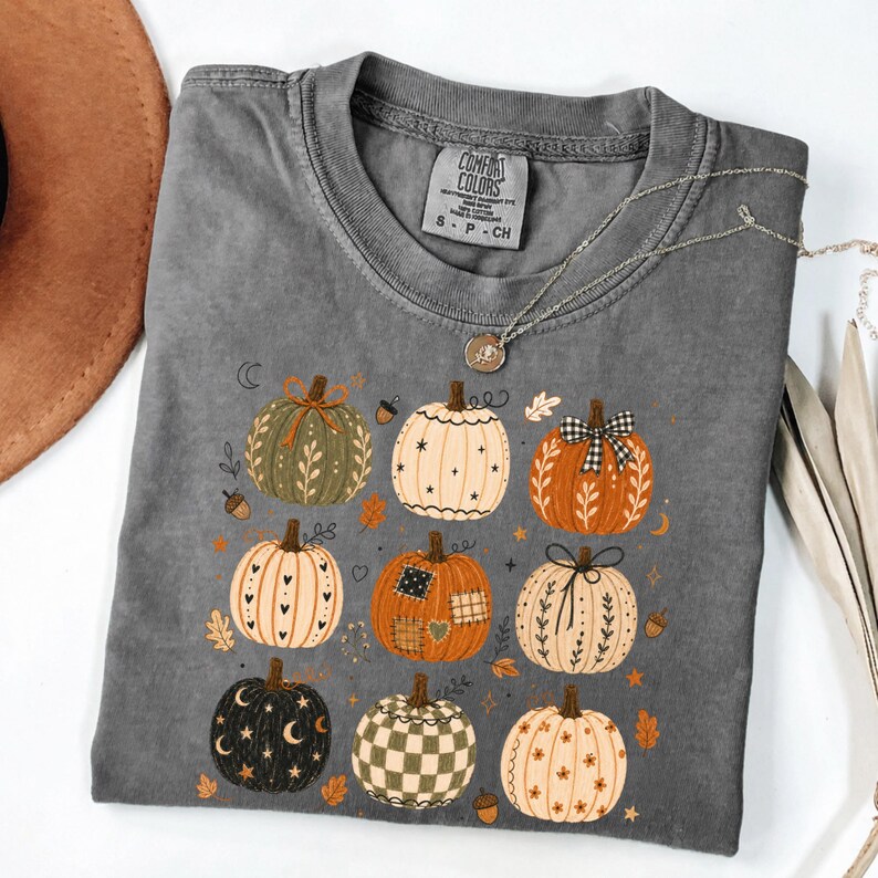 May include: A charcoal gray short-sleeve t-shirt featuring a graphic print of nine pumpkins in various patterns and colors, including orange, green, and cream. The shirt has a crew neckline and a Comfort Colors brand label inside the collar.