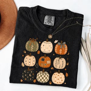 May include: A black short-sleeve t-shirt featuring a graphic print of nine stylized pumpkins in various colors and patterns, including checkered, floral, and heart designs, accented with small stars, moons, and acorns scattered around the pumpkins.