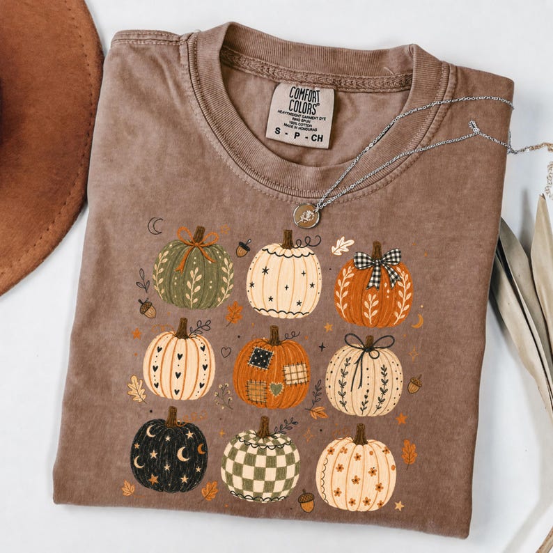 May include: A brown short-sleeve t-shirt featuring a graphic print of nine pumpkins in various colors and patterns, including stars, hearts, and plaid. The shirt has a Comfort Colors brand label at the neck and is paired with a silver chain necklace.