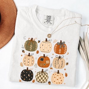 May include: A white short-sleeve t-shirt featuring a graphic print of nine pumpkins in various colors and patterns, including orange, green, black, and cream. The design includes small stars, leaves, acorns, and moons scattered around the pumpkins.
