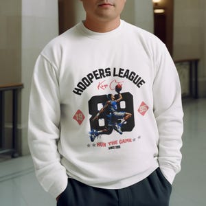 May include: A white long-sleeve sweatshirt featuring a graphic print with the text HOOPERS LEAGUE, Kings City, 89, RUN THE GAME, and SINCE 1995. The design includes a basketball player in a blue uniform jumping to dunk a basketball.