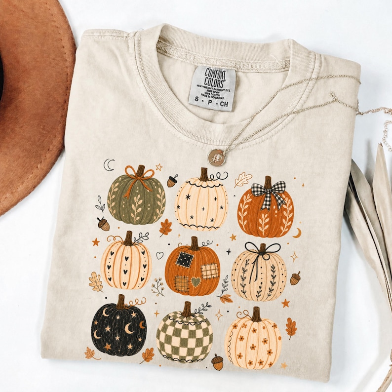 May include: A beige short-sleeve t-shirt with a Comfort Colors label at the neck. The front features a graphic print of nine pumpkins in various colors and patterns, including checkered, floral, and star designs, accented by small leaves and acorns.