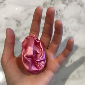 May include: A small, round, pink satin fabric pouch with a gathered top and a knotted closure. The material has a smooth, reflective sheen and is bunched together, creating soft folds and shadows across the surface of the circular object.