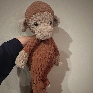 May include: A handmade crochet monkey plush toy crafted from soft, textured brown and beige yarn. The stuffed animal features a rounded head, prominent ears, a distinct snout, and long limbs with contrasting beige hands and feet, all with black eyes.
