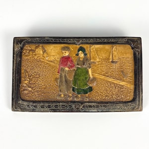 May include: A rectangular metal box with a dark, ornate border. The lid features a relief scene of two figures in traditional clothing, one in a red top and one in a green apron, standing on a cobblestone path near a windmill and sailboats.