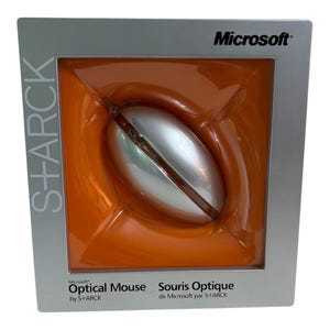 May include: A silver Microsoft optical mouse by S+ARCK encased in a square silver box with an orange interior. The packaging features the text Microsoft, S+ARCK, and Optical Mouse by S+ARCK, Souris Optique de Microsoft par S+ARCK printed on the front.