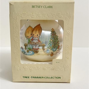 May include: A round, cream-colored Betsey Clark Tree-Trimmer Collection ornament inside a rectangular box. The ornament features an illustration of two children reading a book on a bench near a decorated Christmas tree with gifts underneath.