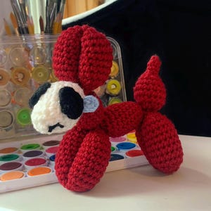 May include: A handmade crochet balloon dog in red, white, black, and light blue. The dog has a white head with black ears and eyes, a red body, and a light blue accent. The dog is sitting on a white surface.