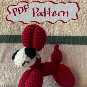 May include: A handmade red crochet dog in a balloon animal style. The dog has a white and black head, with a red body and tail. The text "PDF Pattern" is in a red banner above the dog.
