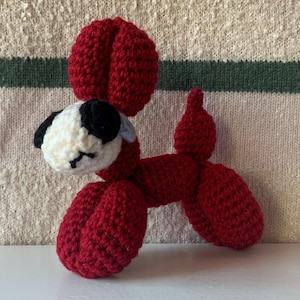 May include: A handmade crochet dog in a balloon animal style. The dog is primarily red with a white and black head, and a small blue accent. The toy is made of yarn and has a whimsical, playful appearance.