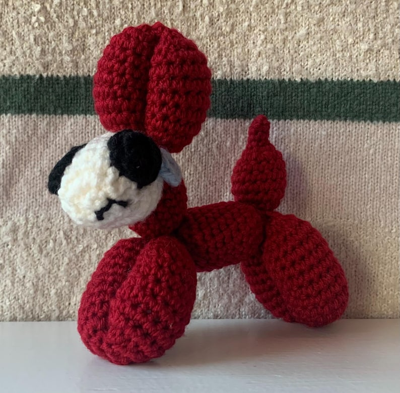 May include: A handmade crochet dog in a balloon animal style. The dog is primarily red with a white and black head, and a small blue accent. The toy is made of yarn and has a whimsical, playful appearance.