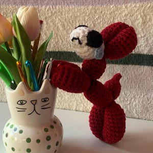 May include: A handmade red crochet dog with a white and black head, positioned next to a ceramic cat-shaped vase filled with tulips and crochet hooks. The vase has green polka dots and a cat face design.