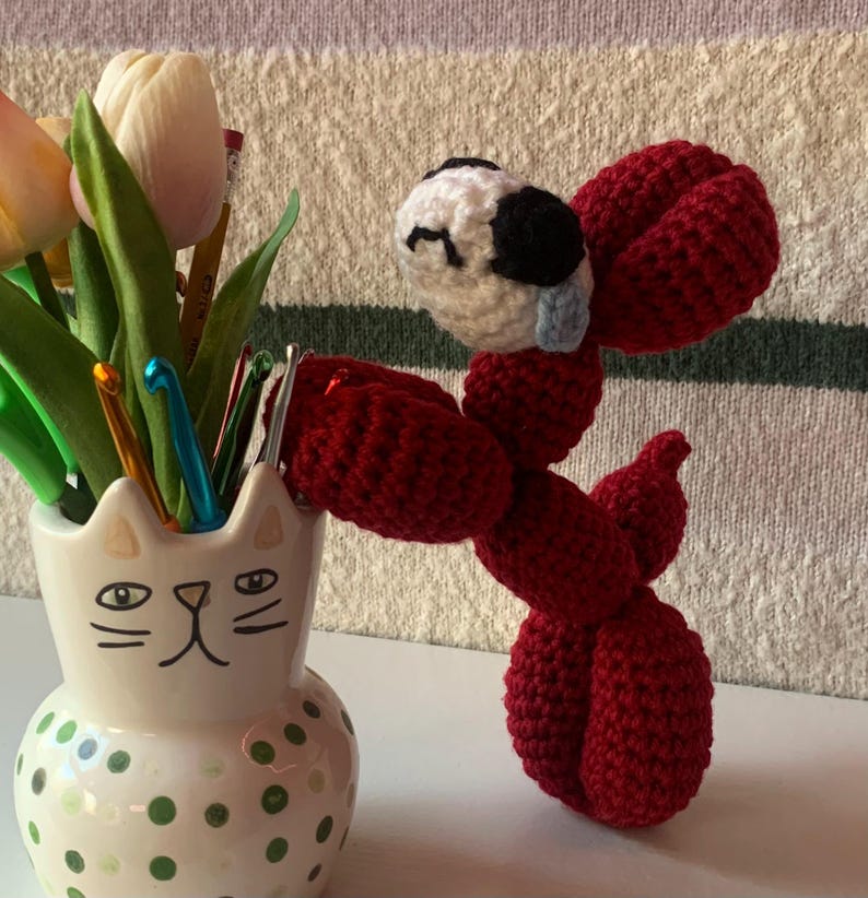 May include: A handmade red crochet dog with a white and black head, positioned next to a ceramic cat-shaped vase filled with tulips and crochet hooks. The vase has green polka dots and a cat face design.
