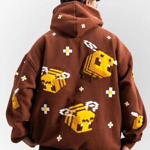 Può includere: Felpa con cappuccio marrone con api pixelate gialle e bianche ricamate e dettagli a croce bianchi. La felpa ha un cappuccio con coulisse e una vestibilità ampia. Il design è sul retro.
