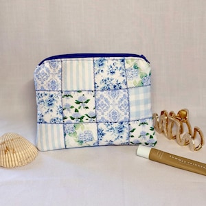 May include: A patchwork pouch with a blue zipper, featuring various blue and white floral and striped patterns. The pouch is rectangular and appears to be made of fabric. A gold-colored hair clip, a seashell, and a tube of Summer Fridays lip balm are also visible.
