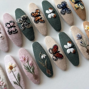 May include: An assortment of embroidered hair clips in various colors. The clips feature detailed floral and butterfly designs in shades of pink, green, beige, and brown. The clips are oval-shaped and made of textured fabric.